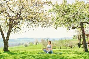 ao ar livre retrato do feliz jovem grávida mulher praticando youga dentro Primavera jardim debaixo florescendo maçã árvores, saudável estilo de vida foto