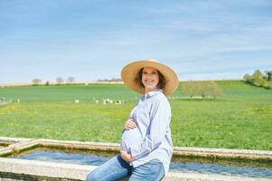 ao ar livre retrato do feliz jovem grávida mulher desfrutando agradável dia dentro interior, sentado em rega fonte, verde pasto com vacas em fundo foto