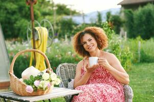 ao ar livre retrato do lindo maduro mulher em repouso dentro verão jardim, sentado dentro acolhedor cadeira, segurando copo do chá ou café, cesta com recentemente cortar Rosses em pé em a mesa foto