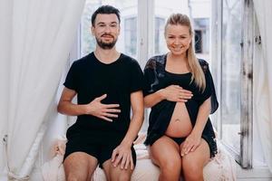casal engraçado feliz posando na janela e mostrando a barriga da mulher grávida.
