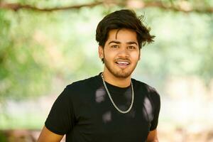 sorridente jovem indiano homem retrato dentro Preto t camisa e prata pescoço cadeia ao ar livre verde público parque foto