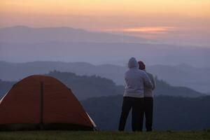 casal é abraçando cada de outros de a barraca durante durante a noite acampamento enquanto olhando às a lindo cênico pôr do sol sobre a montanha para ao ar livre aventura período de férias viagem conceito foto