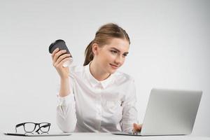 mulher de negócios sorridente, sentada e trabalhando atrás de um laptop, segurando uma xícara de café e olhando para a tela foto