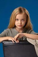 pequeno menina com cinzento olhos e loiro cabelo senta em uma cadeira foto