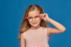 elegante pequeno menina dentro uma Rosa vestir e óculos é em pé em uma azul fundo. foto