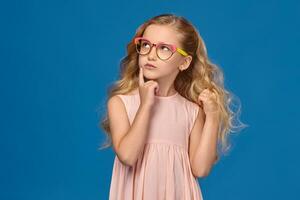 elegante pequeno menina dentro uma Rosa vestir e óculos é em pé em uma azul fundo. foto