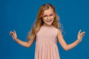 elegante pequeno menina dentro uma Rosa vestir e óculos é em pé em uma azul fundo. foto