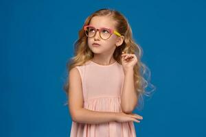 elegante pequeno menina dentro uma Rosa vestir e óculos é em pé em uma azul fundo. foto