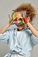 lindo pequeno menina com uma pintado mãos e bochechas é posando em uma cinzento fundo. foto