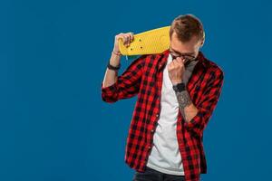carismático alegre jovem barbudo homem vestido dentro xadrez camisa, branco camiseta e copos, com amarelo skate dentro dele mãos. estúdio tiro com azul fundo foto