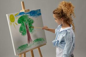 encantador escola menina é pintura com uma aguarela escova em a cavalete, em pé em uma cinzento fundo. foto