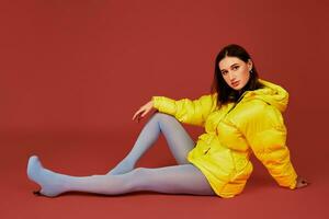 estúdio retrato do jovem morena mulher sentado em a chão dentro amarelo baixa Jaqueta e cinzento azul calcinha mangueiras ou meias foto