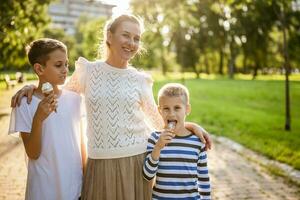 retrato do feliz solteiro mãe com dela dois filhos dentro parque em ensolarado dia. Rapazes estão comendo gelo creme. foto