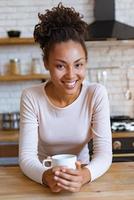 menina bonita se senta à mesa com uma xícara de chá ou café da manhã e sorrindo, olhando para a câmera foto