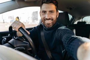 ótima selfie. bonito homem barbudo segurando a câmera e fazendo selfie enquanto está sentado no carro no banco do motorista e olhando para a câmera com um sorriso de prazer foto