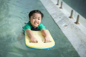 ásia criança criança menina sorridente engraçado vestindo natação terno usando pontapé borda para natação ou inclinado Treinamento em natação piscina dentro água parque. água atividade para crianças em verão feriado. foto