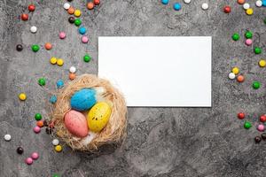 sobre um fundo de gesso cinza encontra-se um ninho de pássaro com ovos coloridos, uma folha em branco, um cartão de páscoa e doces foto