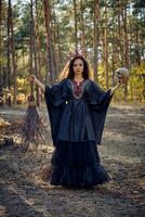 bruxa dentro preto, grandes vestir, com vermelho coroa dentro dela grandes cabelo. posando com vassoura e crânio dentro pinho floresta. feitiços, Magia e feitiçaria. cheio comprimento. foto