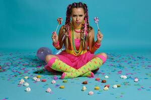 adorável menina com uma multicolorido tranças Penteado e brilhante inventar, posando dentro estúdio com pirulito, ar balões e confete contra uma azul fundo. foto