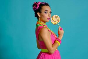 adorável menina com uma multicolorido tranças Penteado e brilhante inventar, posando dentro estúdio contra uma azul fundo, segurando uma pirulito dentro dela mão. foto