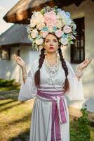 morena menina dentro uma branco ucraniano autêntico nacional traje e uma guirlanda do flores é posando contra uma branco cabana. fechar-se. foto