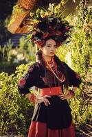 morena menina dentro Preto e vermelho bordado ucraniano autêntico nacional traje e uma guirlanda do flores é posando em pé contra uma branco cabana. foto