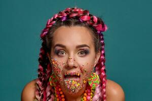 adorável menina com uma multicolorido tranças Penteado e brilhante inventar, posando dentro estúdio contra uma azul fundo. colorida cobertura é em dela face. foto