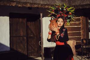 morena menina dentro uma Preto e vermelho ucraniano bordado autêntico nacional traje e uma guirlanda do flores é posando em pé às a portão. foto