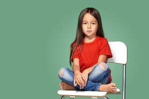 pequeno menina vestindo vermelho t-curto e posando em cadeira foto