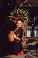 morena menina dentro uma Preto e vermelho ucraniano bordado autêntico nacional traje e uma guirlanda do flores é posando segurando uma jarro e em pé às a portão. foto