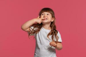 fechar-se retrato do uma pequeno morena menina vestido dentro uma branco camiseta posando contra uma Rosa estúdio fundo. sincero emoções conceito. foto