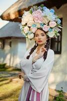 morena menina dentro uma branco ucraniano autêntico nacional traje e uma guirlanda do flores é posando contra uma branco cabana. fechar-se. foto