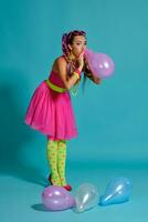 adorável menina com uma multicolorido tranças Penteado e brilhante inventar, posando dentro estúdio com ar balões contra uma azul fundo. foto