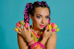 adorável menina com multicolorido tranças Penteado e brilhante inventar, segurando macarons entre dela dedos, posando dentro estúdio contra uma azul fundo. foto