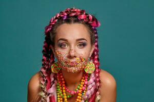 adorável menina com uma multicolorido tranças Penteado e brilhante inventar, posando dentro estúdio contra uma azul fundo. colorida cobertura é em dela face. foto