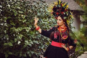 morena menina dentro Preto e vermelho bordado ucraniano autêntico nacional traje e guirlanda do flores é caminhando sozinho e posando dentro uma verde jardim. foto
