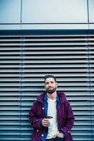 ao ar livre outono ou inverno retrato do bonito hipster homem com barba, branco camiseta, azul camisa e marrom Jaqueta segurando copo do quente café. foto