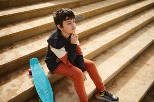 pensativo multi étnico Adolescência estudante dentro roupa de esporte, sentado com dele skate em a passos ao ar livre, sonhadoramente longe foto