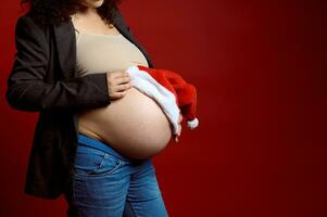 fechar-se barriga do gravídico mulher, expectante mãe segurando santa chapéu em dela grávida barriga, isolado em vermelho estúdio fundo foto