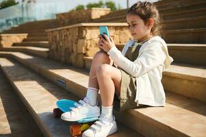 pequeno menina com skate, sentado em passos ao ar livre, usando Smartphone, Verificações social meios de comunicação contente, relógios desenhos animados foto