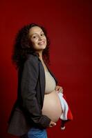 vertical estúdio retrato do uma rindo feliz jovem adulto expectante mãe tocante dela grávida barriga em vermelho fundo foto