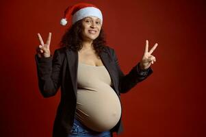 lindo autêntico grávida mulher dentro santa chapéu, sorrisos às Câmera, mostra Paz sinal, expressando positivo emoções foto