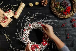 topo Visão do fêmea mãos faço uma Natal guirlanda. embalado presentes e pergaminhos, abeto galhos e Ferramentas em de madeira mesa. foto