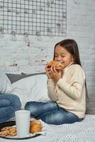 bonita pequeno menina 6 anos velho dentro uma branco suéter e jeans. criança dentro a quarto com uma cama, comendo croissant e bebendo chá ou cacau. foto