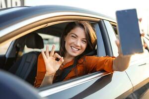 carro alugando aplicativo. feliz fêmea mostrando em branco Smartphone enquanto sentado dentro do auto, sorridente mulher demonstrando cópia de espaço para Móvel propaganda ou local na rede Internet, brincar foto