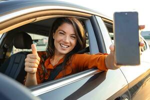 carro alugando aplicativo. feliz fêmea mostrando em branco Smartphone enquanto sentado dentro do auto, sorridente mulher demonstrando cópia de espaço para Móvel propaganda ou local na rede Internet, brincar foto