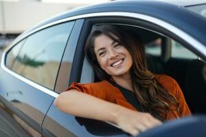 jovem mulher sentada em um carro com a mão para fora da janela. mulher feliz dirigindo um carro e sorrindo. retrato do carro de direção do motorista feminino feliz com cinto de segurança. linda jovem feliz dirigindo o carro. foto