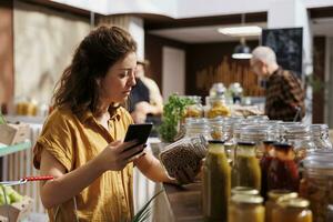 mulher dentro zero desperdício loja usando Smartphone notas aplicativo para Verifica compras lista. meticuloso cliente olhando para reabastecimento despensa às casa com orgânico produtos químicos livre Comida Essenciais foto