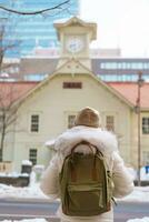 mulher turista visitando dentro sapporo, viajante dentro suéter passeios turísticos sapporo relógio torre com neve dentro inverno. ponto de referência e popular para atrações dentro Hokkaido, Japão. viagem e período de férias conceito foto