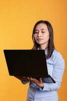 ásia jovem adulto usando computador portátil computador dentro frente do Câmera sobre amarelo fundo, segurando pc e navegando em formação em Internet. morena empresária olhando às dispositivo tela dentro estúdio. foto